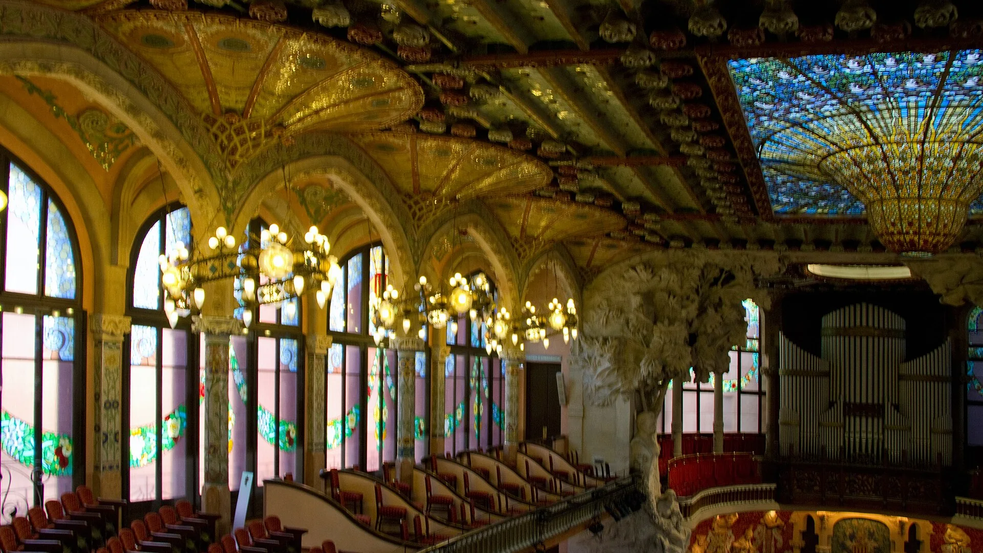 La sala de conciertos del Palau de la Música Catalana vista de lado: ventanales arqueados de vidrieras, lámparas colgantes, un lucernario de vidriera en oro y azul, y el escenario con sus musas esculpidas y su órgano.