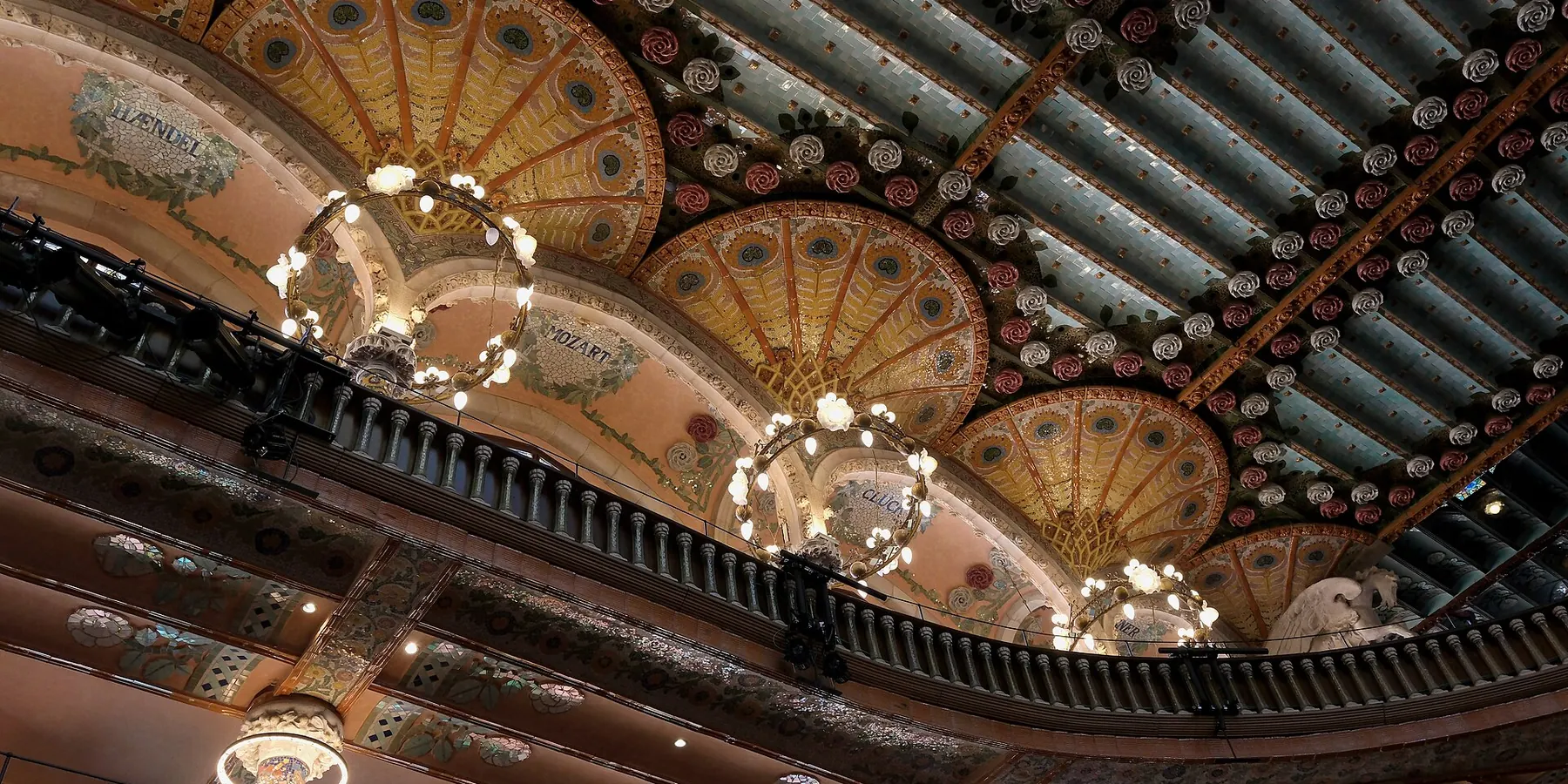 El techo de la sala de conciertos del Palau con ornamentación dorada y verde en forma de abanico, lámparas colgantes en forma de anillo y un balcón curvo debajo.