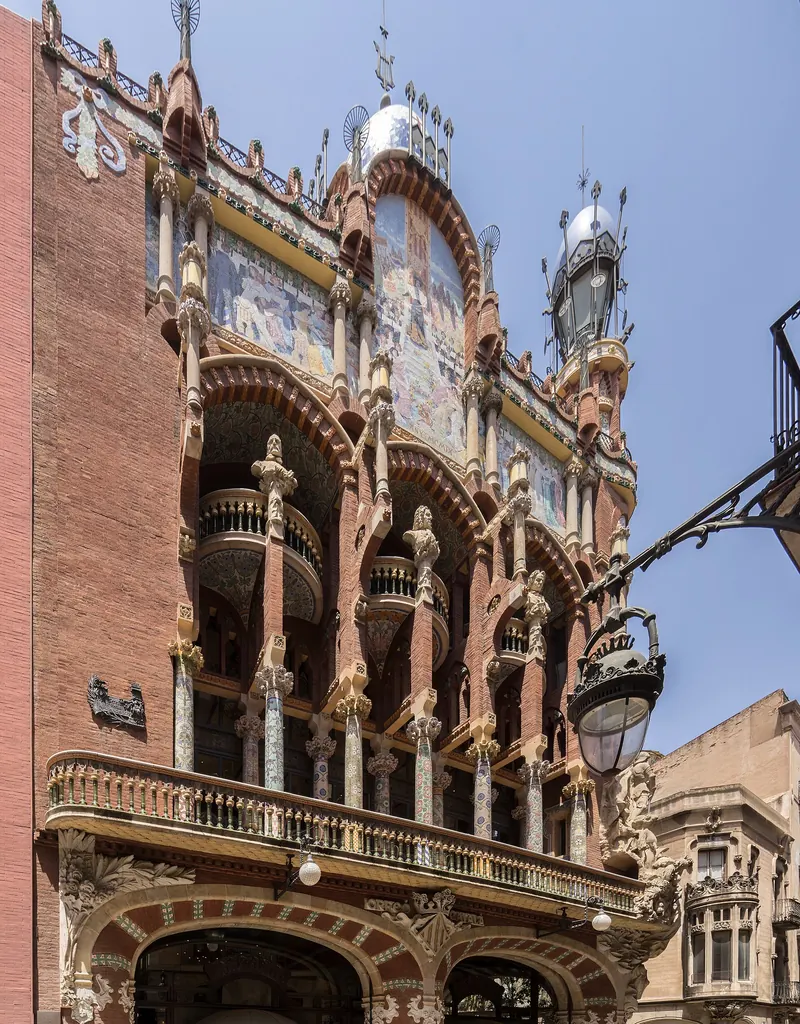 El chaflán del Palau de la Música Catalana en ladrillo rojo, con columnas de mosaico, balcones y un frontón esculpido contra un cielo azul.