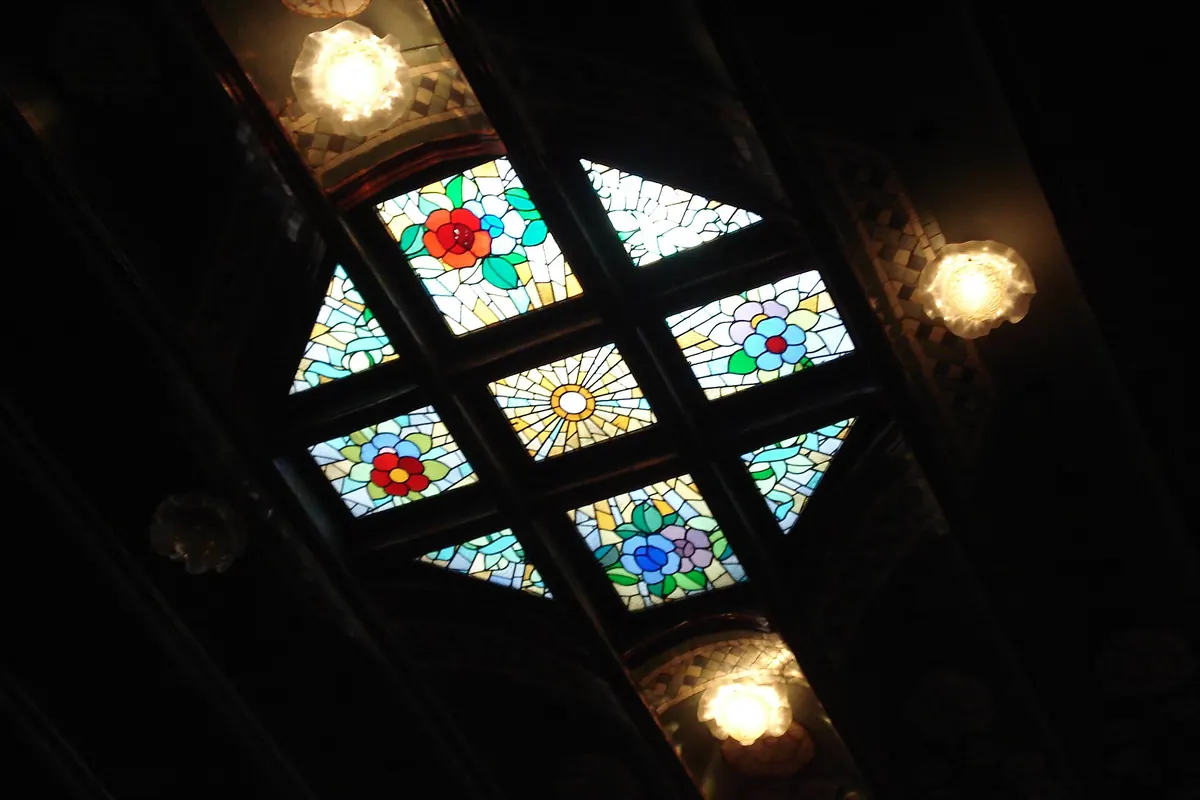 Un pequeño panel de vidriera en el techo del Palau con flores rojas, azules y doradas dispuestas en un patrón de rombos entre dos lámparas.