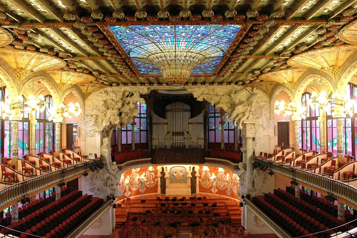 La sala de conciertos del Palau de la Música Catalana desde el patio de butacas, con una vidriera cenital en azul y oro sobre la cabeza y el escenario con sus musas al frente.