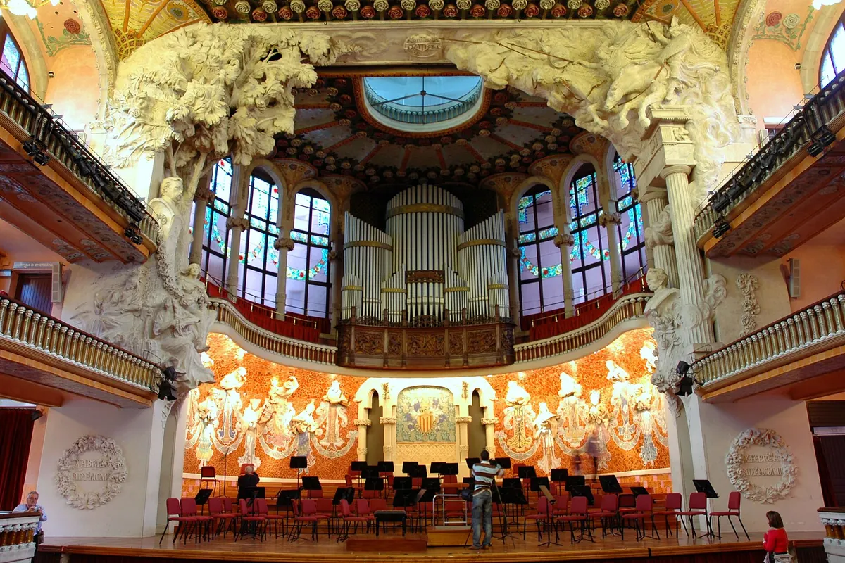 El escenario de la sala de conciertos del Palau con árboles escultóricos blancos, el órgano arriba y un muro de mosaico de musas tras las sillas de una orquesta.